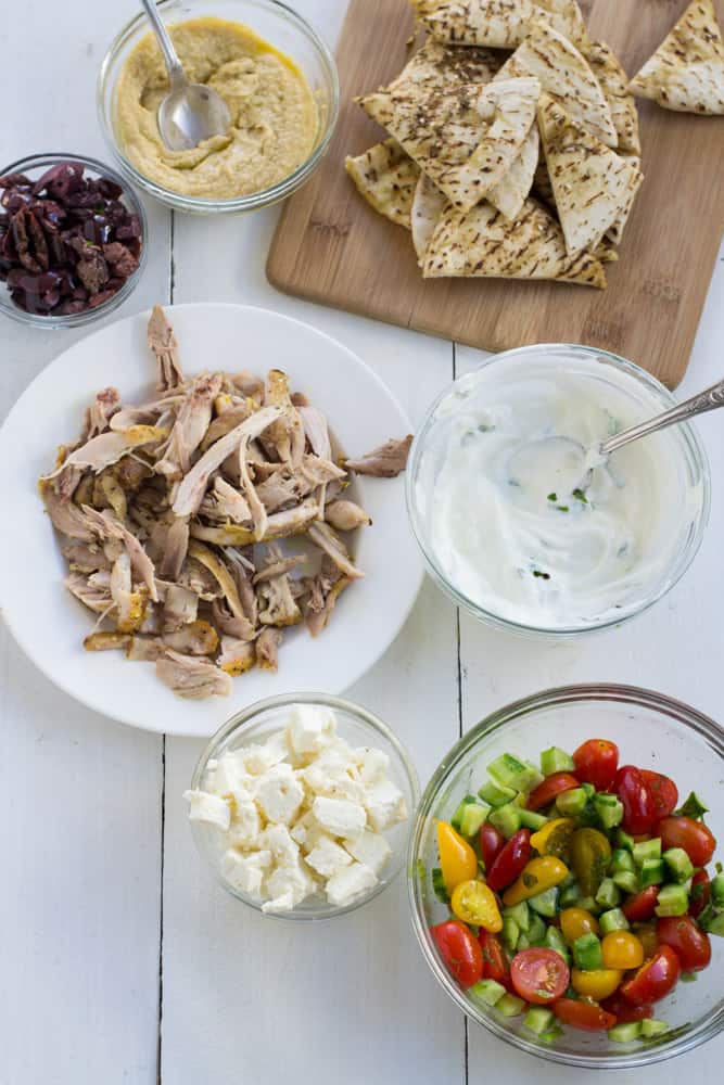 Ingredients for Mediterranean nachos include pita bread cut into wedges, hummus, chopped olives, yogurt mint sauce, shredded roasted chicken and chopped salad.
