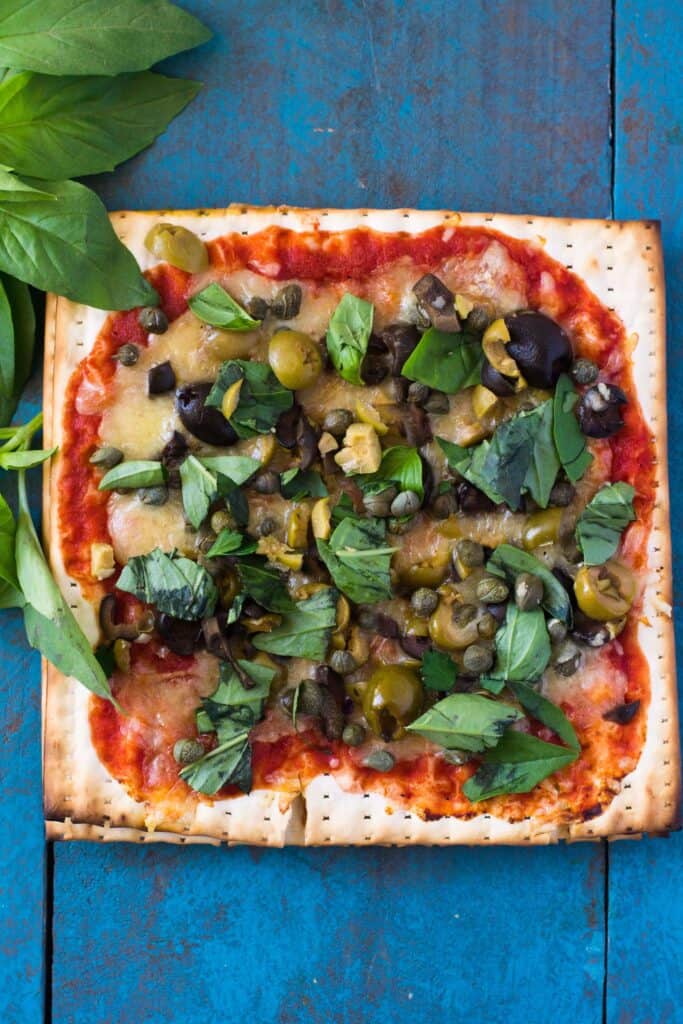 A thin layer of marinara is spread over the matzo and then topped with chopped olives, briny capers and shredded mozzarella. After baked, garnish with a few leaves of fresh basil to brighten up all the savory flavors.