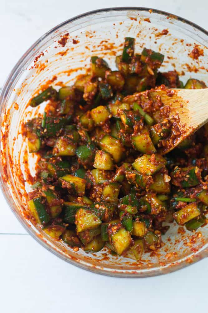 Cucumber kimchi is mixed together in a  large glass bowl. The cucumbers are chopped into ½ in pieces and mixed with Korean red pepper, fresh green onion, grated garlic, grated ginger and sesame seeds. 