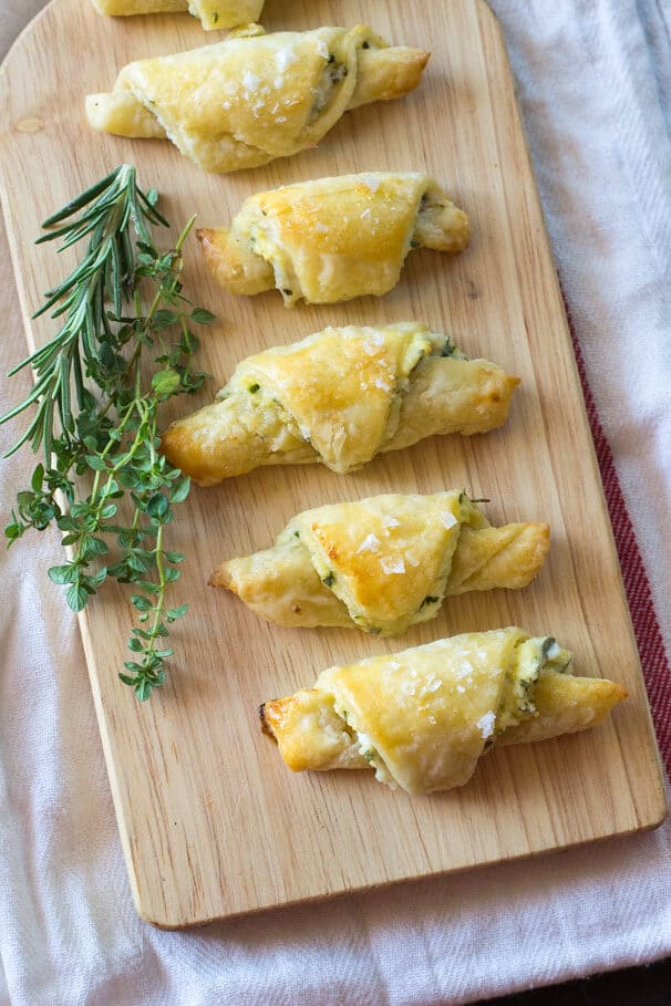 Savory rugelach are lined up on a wooden cutting board with fresh herbs on the side. Rugelach are cookies and these savory versions are seasoned with fresh herbs and Parmesan cheese and filled with a ricotta mixture that is mixed with roasted garlic. 