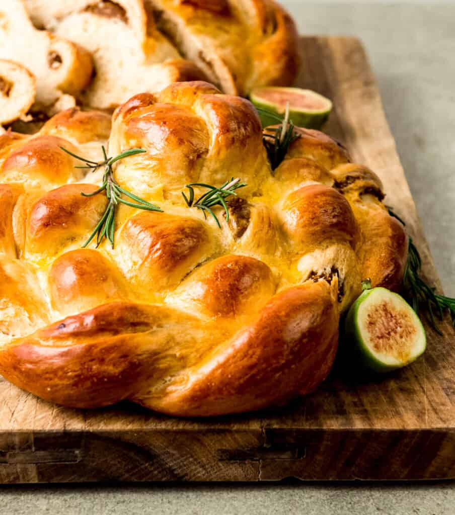Round fig challah bread is garnished with fresh rosemary and fresh figs, illustrating the filling. The filling is stuffed inside the challah strands.