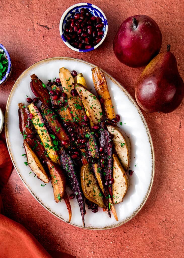 Roasted carrots and pears are seasoned with maple syrup, allspice, cinnamon and pomegranate molasses until caramelized and once roasted are arranged on a wide platter. They are garnished with pomegranate seeds and fresh chopped parsley. Around the platter is whole red anjou pears, a bowl of pomegranate seeds and a bowl of fresh chopped herbs.