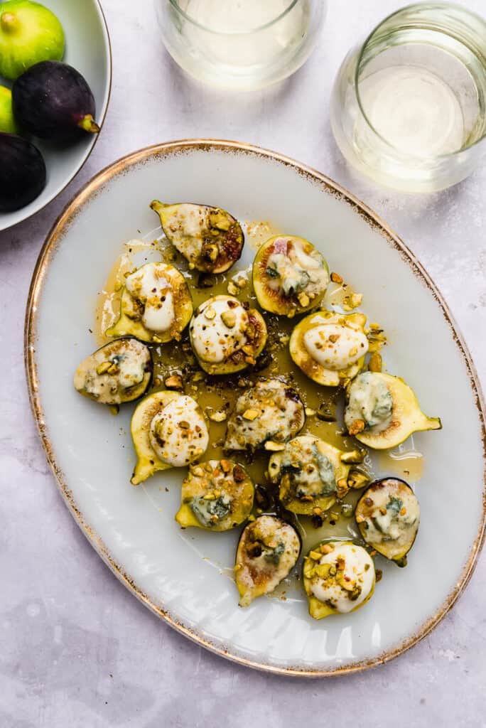 Fresh figs are cut in half and layed out on a platter. Some figs are topped with crumbled blue cheese and some with whipped goat cheese and garnished with chopped pistachios and honey.