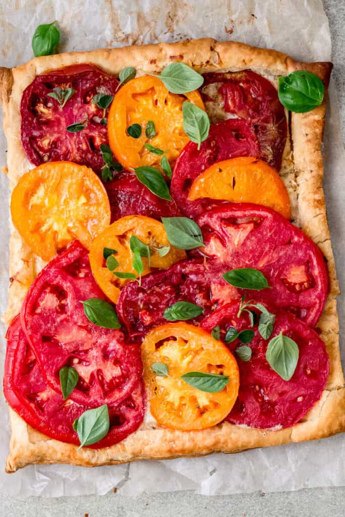 Colorful slices of heirloom tomatoes are layered on top of cooked puff pastry with a thin layer of mascarpone cheese under the tomatoes. The mascarpone tart is garnished with a few leaves of fresh basil. 