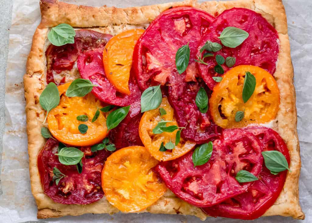 Slices of yellow and red heirloom tomatoes are layered on top of cooked puff pastry with a spread of mascarpone cheese in between. The mascarpone tart is topped with leaves of fresh basil.