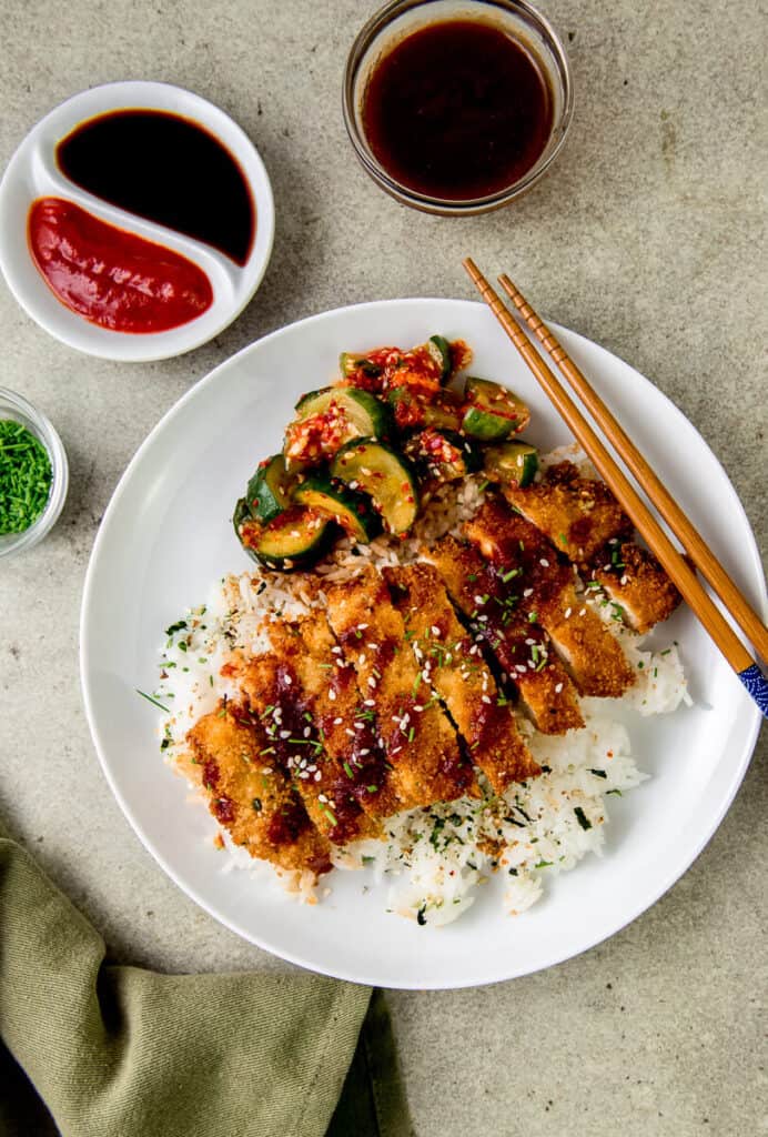 Chicken katsu is a fried chicken cutlet thats coated with panko breadcrumbs and fried to golden brown. The fried chicken is served on top of white rice with katsu sauce drizzled on top and cucumber kimchi on the side. 