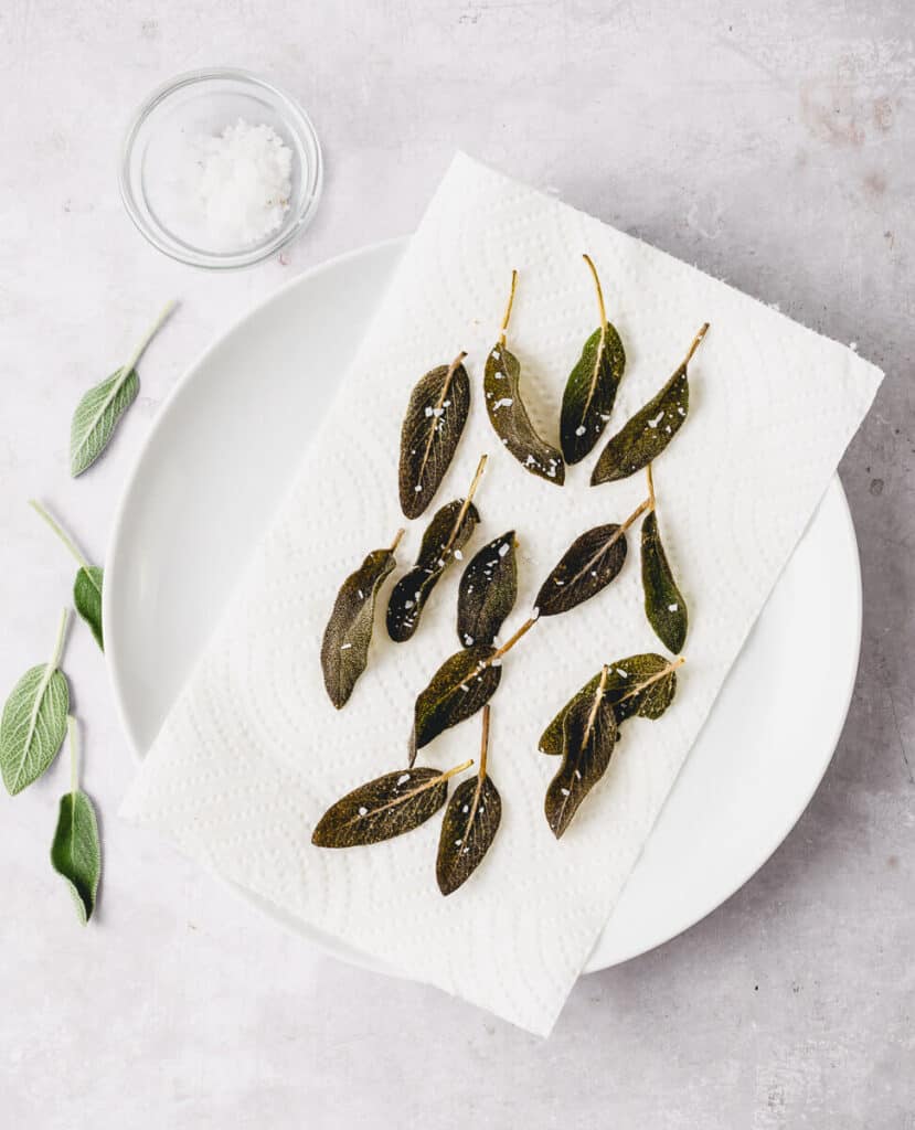 Fried sage leaves are on a paper towel lined plate and sprinkled with flakey sea salt. 