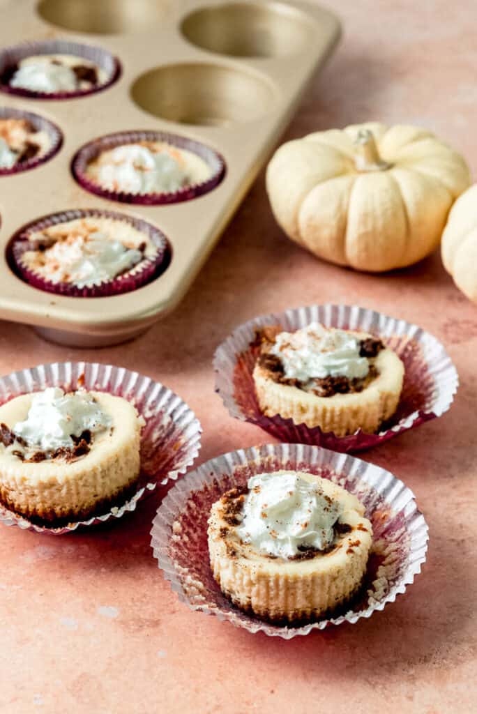 Three mini cheesecakes are displayed in cupcake liners and next to a cupcake tin. The individual sized cheesecakes are swirled with pumpkin and garnished with whipped cream and grated nutmeg. 