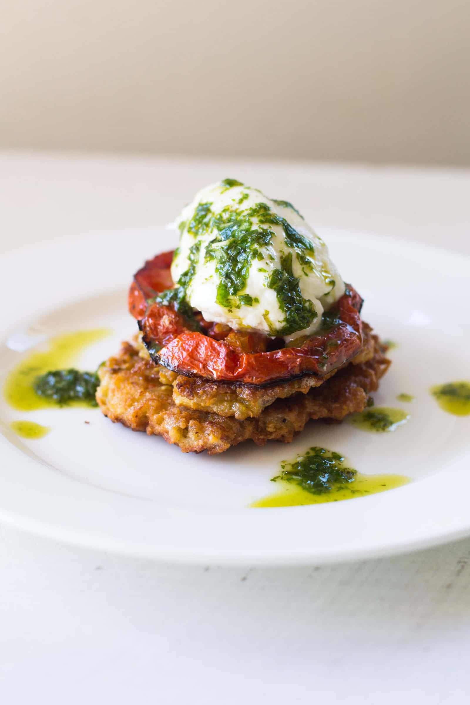 Two potato latkes stacked with slices of roasted tomato and topped with burrata cheese and basil oil.
