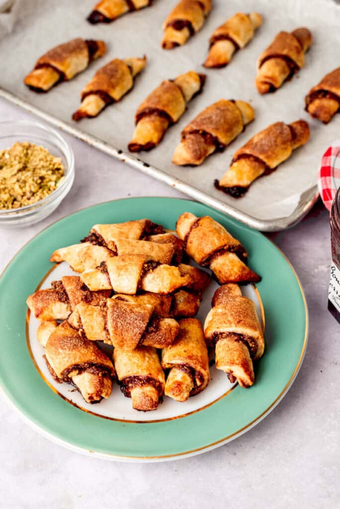Cherry pistachio rugelach are piled on a plate with more rugelach on a cookie sheet behind the plate.