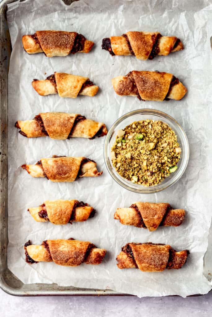 Cookie sheet with baked cherry pistachio rugelach and a small bowl of chopped pistachios.