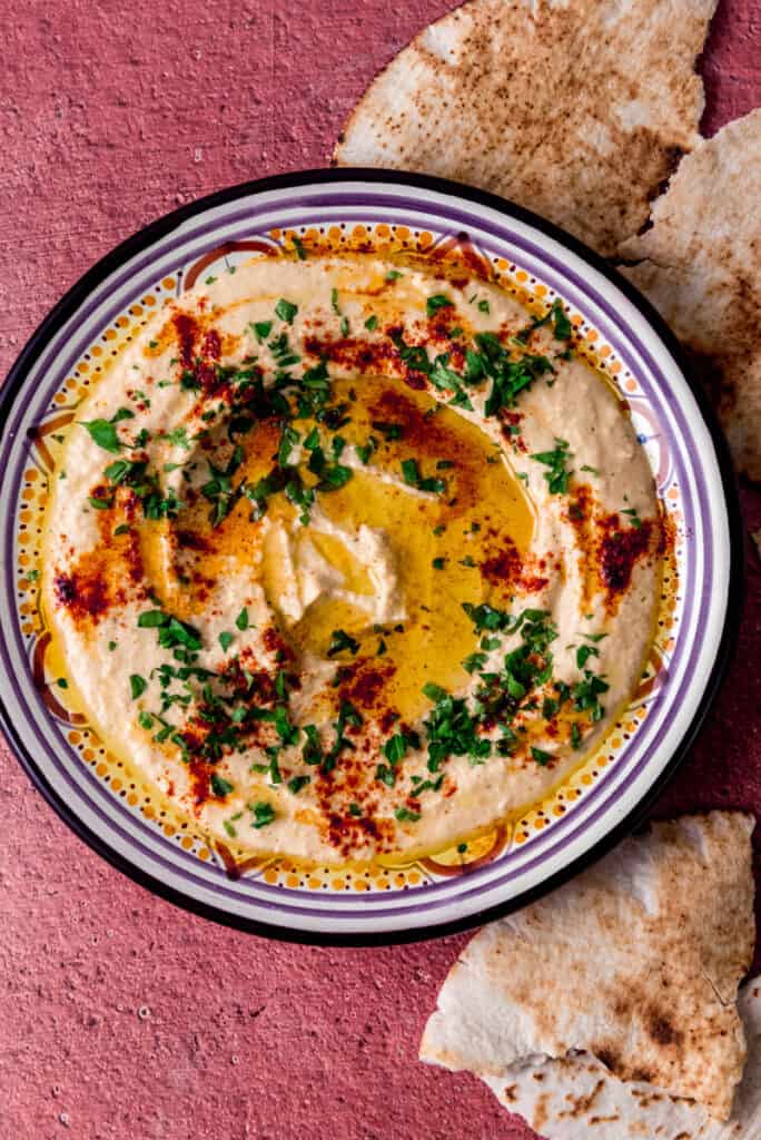 Homemade chickpea hummus is spread onto a round plate ad garnished with olive oil, fresh herbs and paprika and served with torn pita bread.