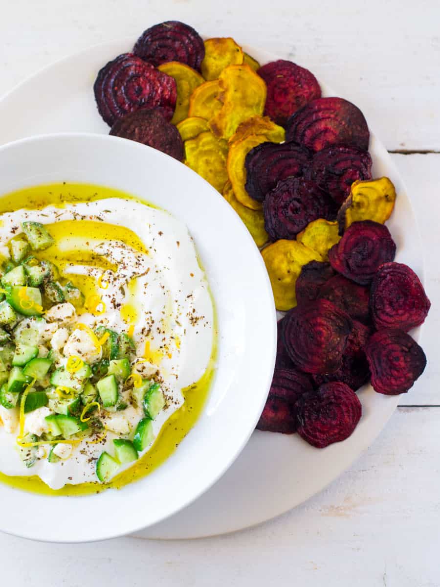 Red and yellow baked beet chips are served alongside a creamy labneh dip that is garnished with cucumber and lemon zest.