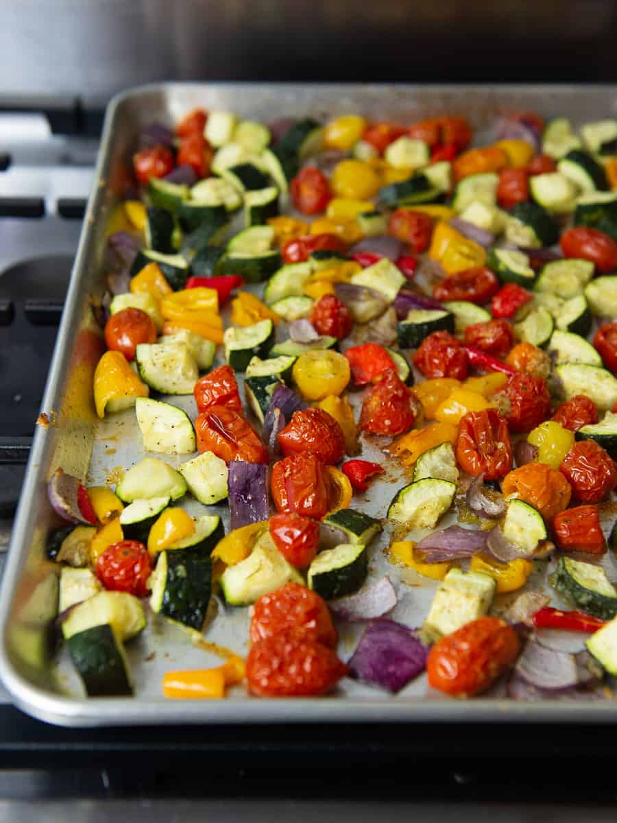 Roasted vegetables on a baking sheet, including cherry tomatoes, cubed zucchini, red onion and bell peppers.