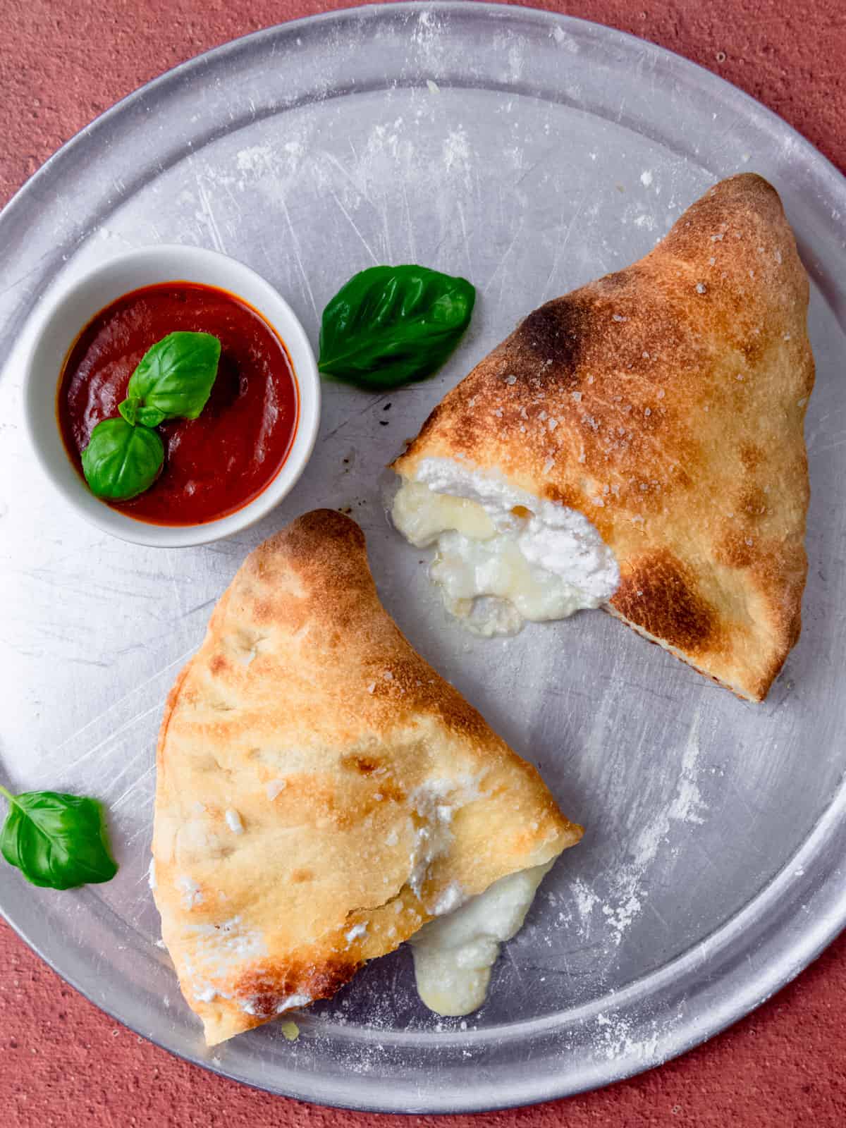 Baked cheese calzone tat is cut in half and served with a bowl of marinara sauce for dipping.