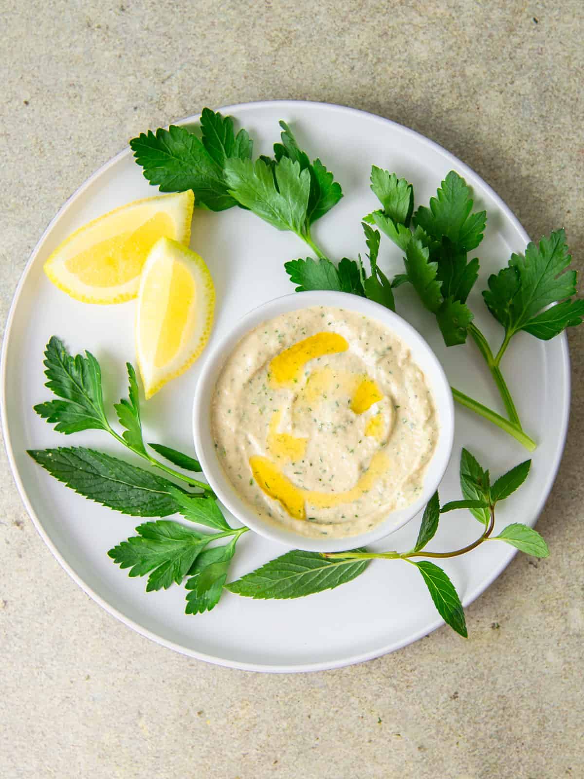 Lemon herb tahini sauce in a bowl with fresh herbs scattered around.